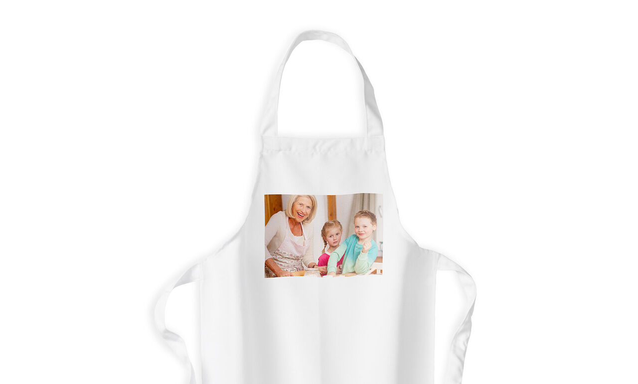En vit förkläde med ett glatt familjefoto av en äldre kvinna och två barn som har roligt i köket.