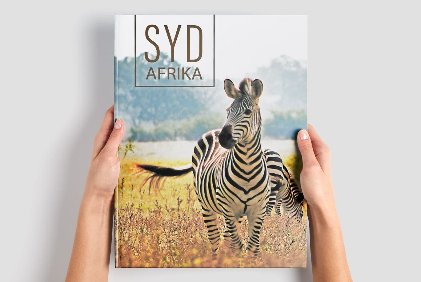 En person håller en fotobok med ett zebramotiv på omslaget. Bakgrunden visar ett afrikanskt landskap.
