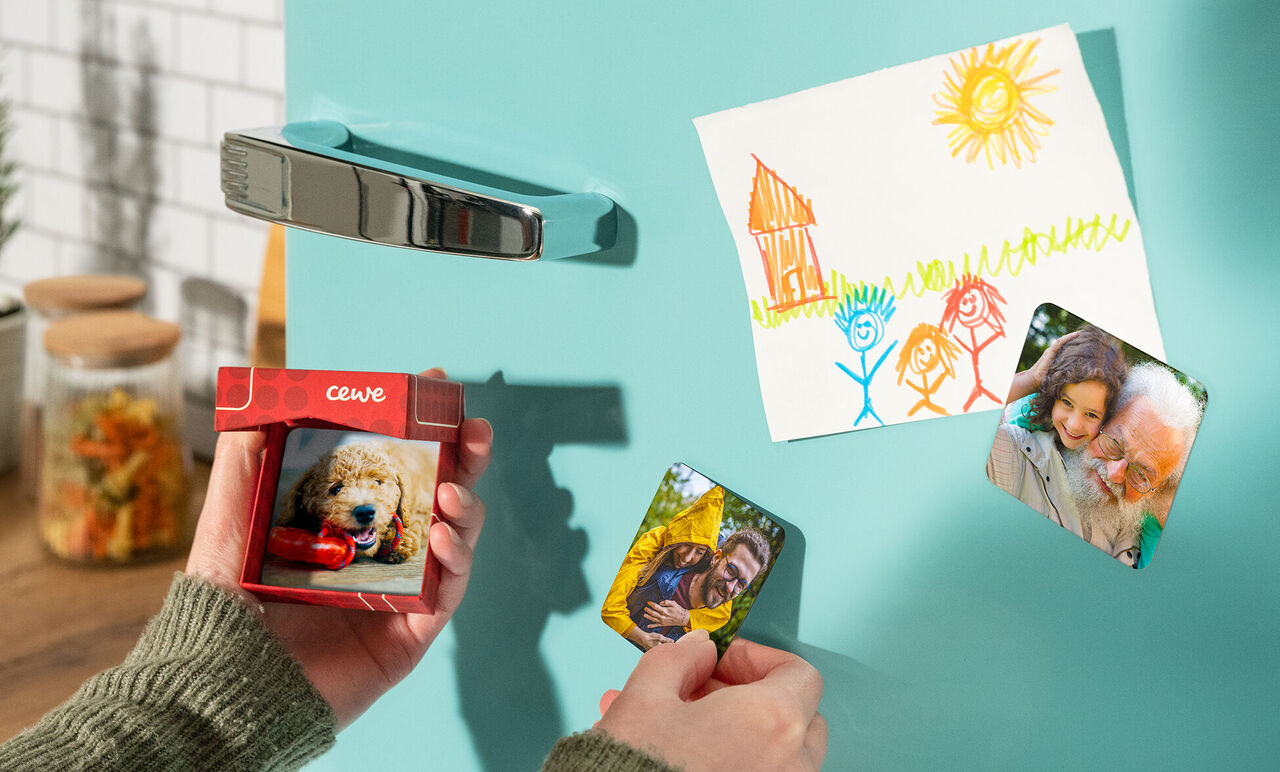 En hand håller ett litet CEWE kort med hundbild, medan en teckning hänger på kylskåpet.
