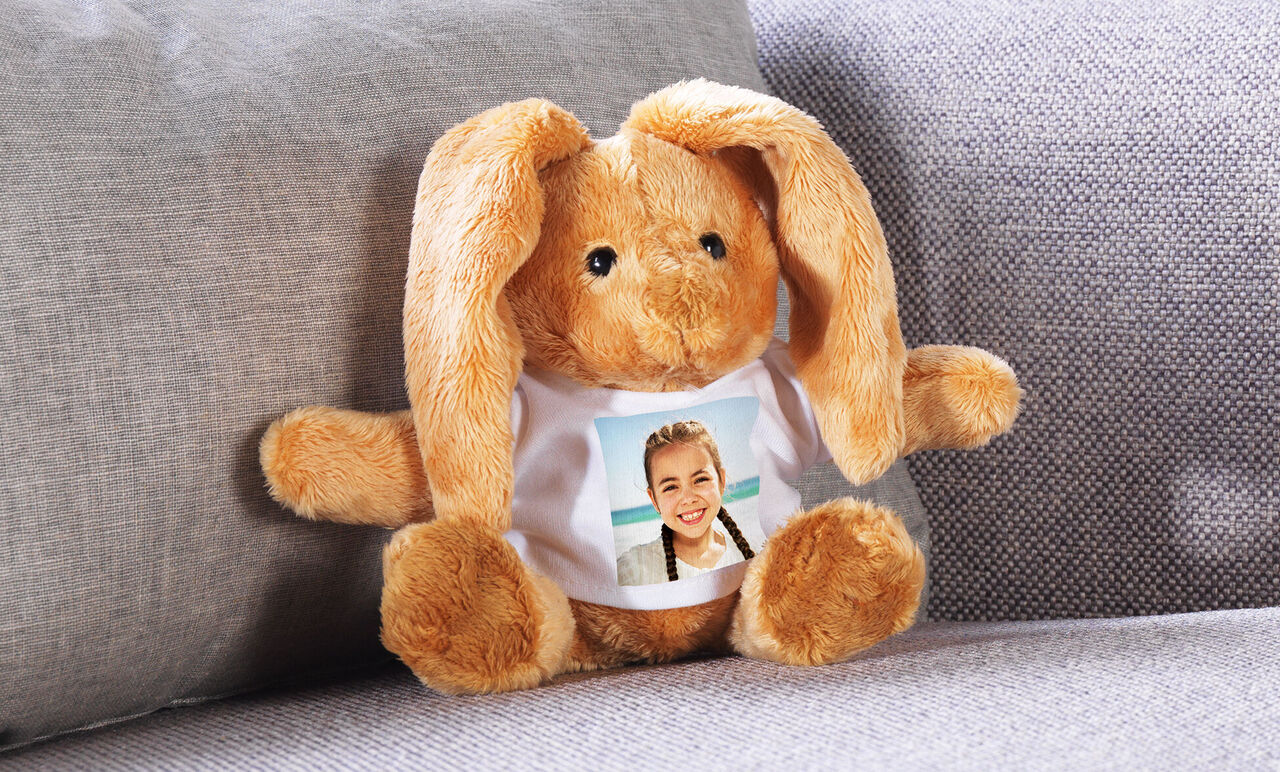 En söt plyschkanin sitter på en soffa. Den bär en t-shirt med foto.