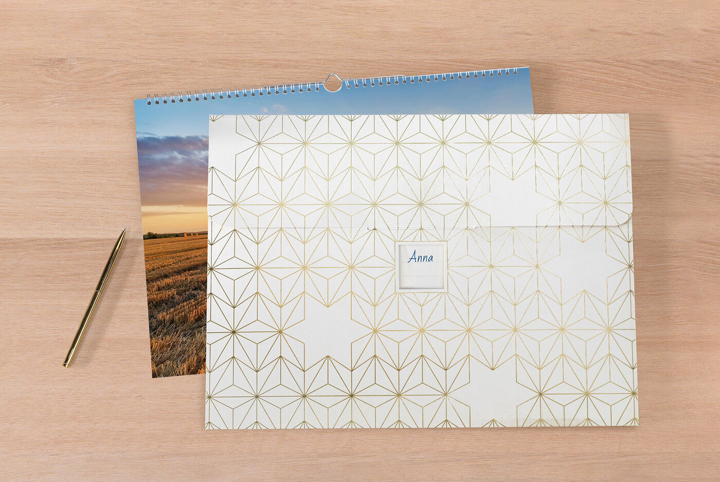 En kärleksfullt designad CEWE fotokalender på ett trädligt bord, bredvid ett dekorativt kuvert med namnet 'Anna' och en gyllene penna.