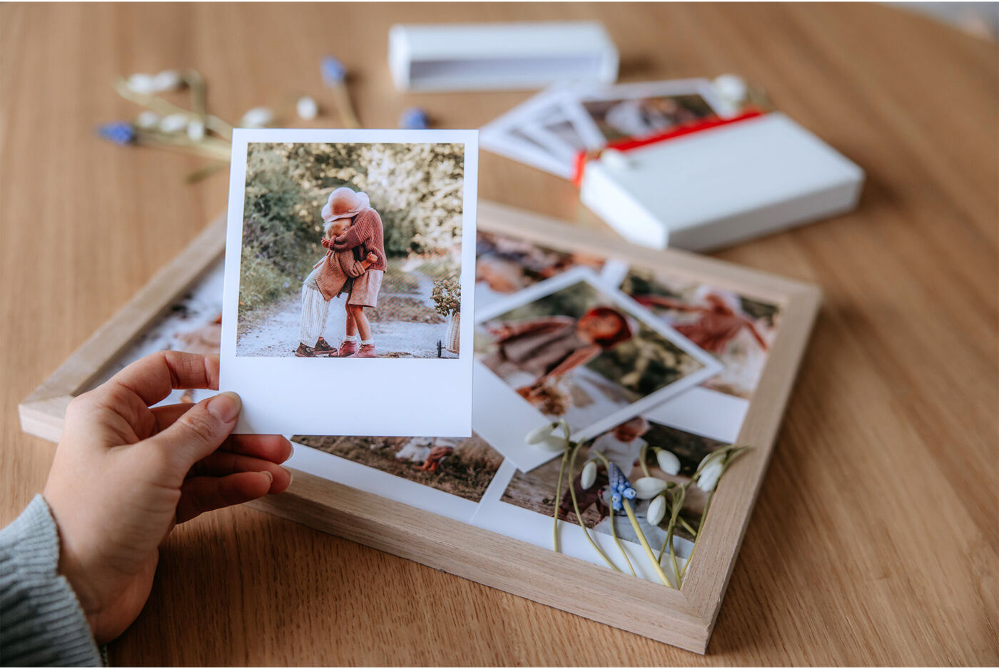 En hand håller ett Polaroidfoto av två barn som kramar varandra, med fler foton i bakgrunden.