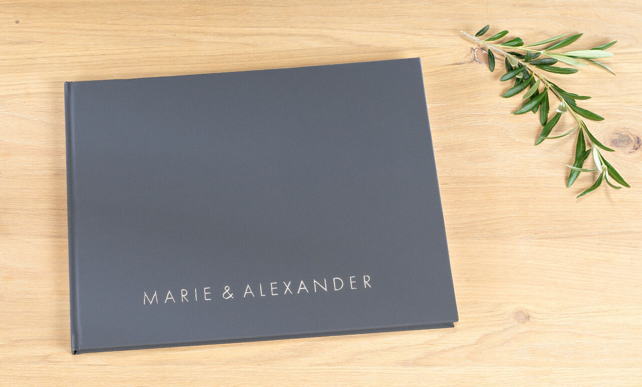 Mörk fotobok med titeln "Marie & Alexander" på ett träunderlag med olivkvist.