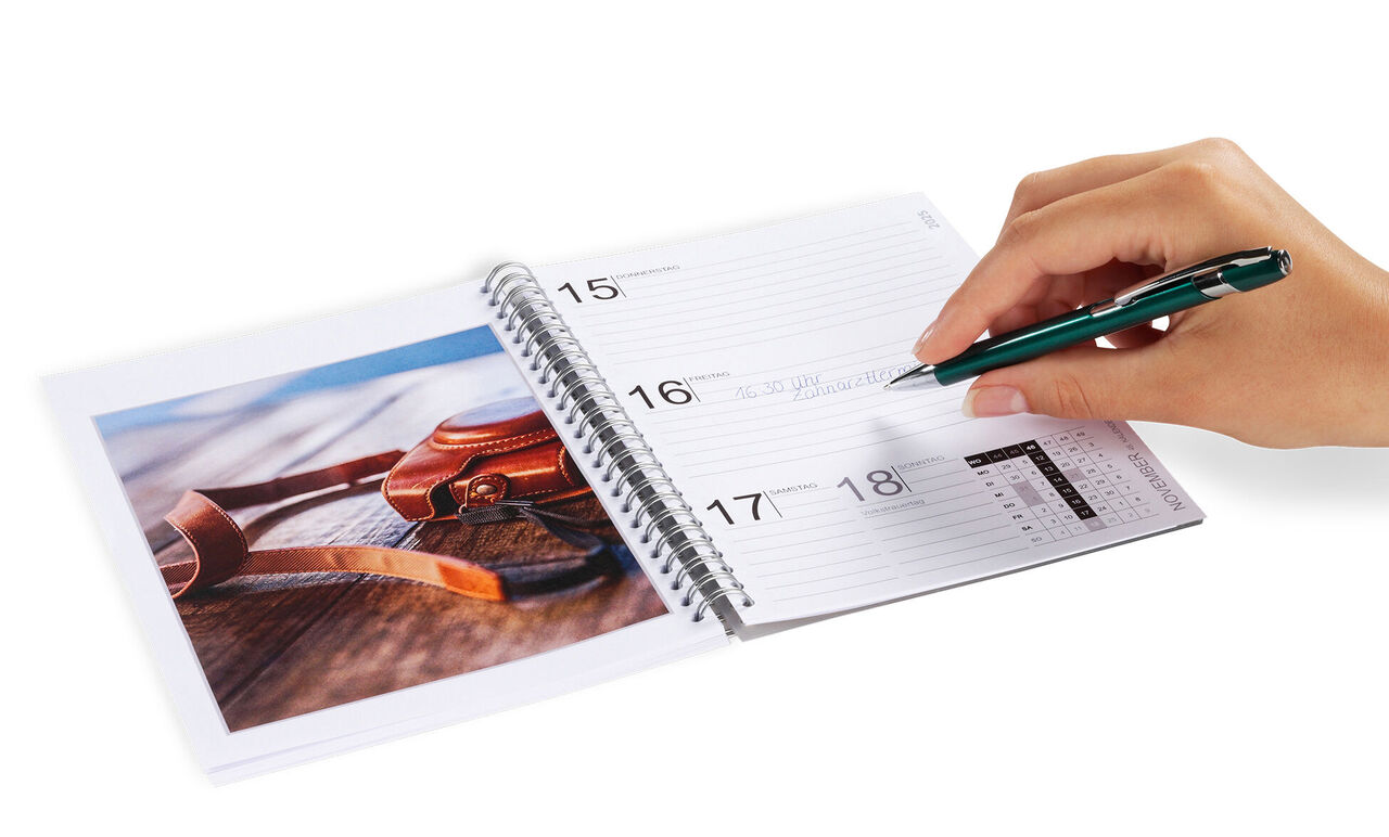 En hand skriver i en kalender, bredvid ligger ett foto med solglasögon på ett bord.