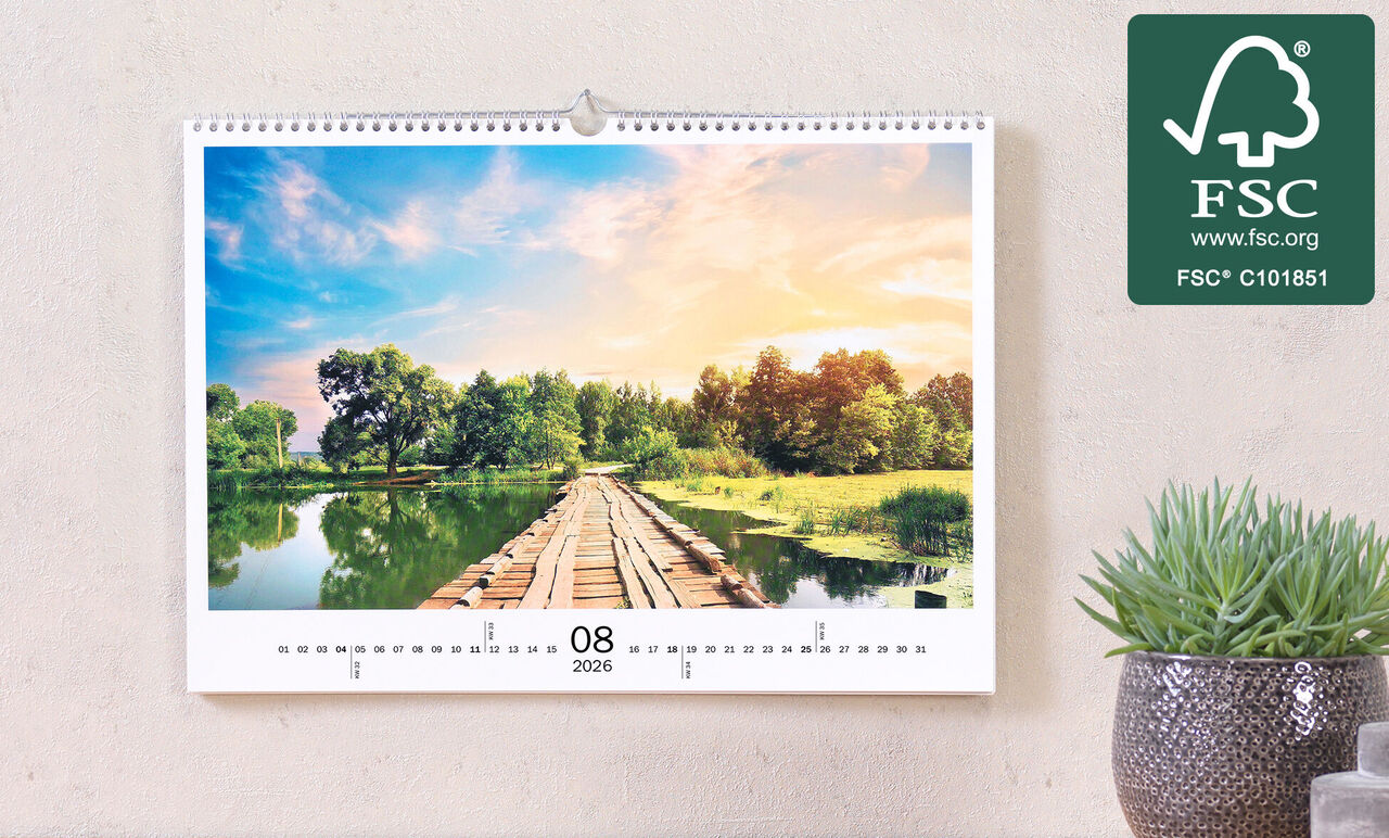 Hängande fotokalender visar en pittoresk landskap med en träbro över en lugn vattenyta, omgiven av frodig grönska.