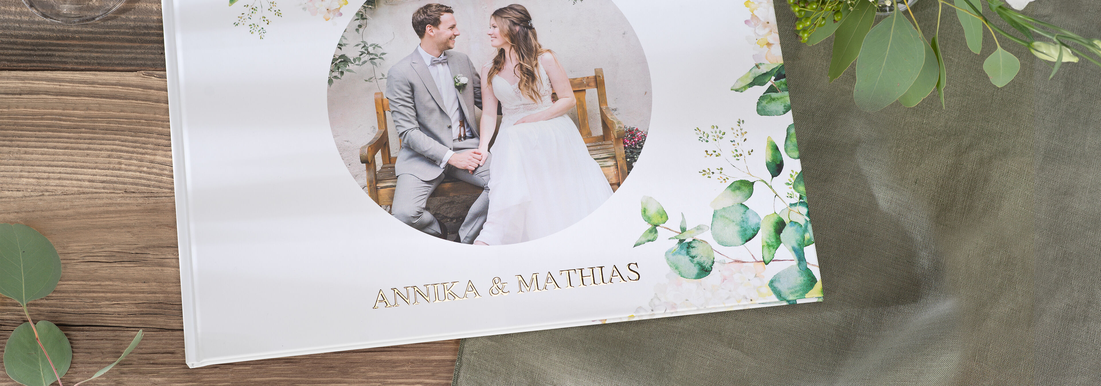 En vacker CEWE fotobok med titeln Annika & Matthias på ett träbord, omgiven av gröna blad.