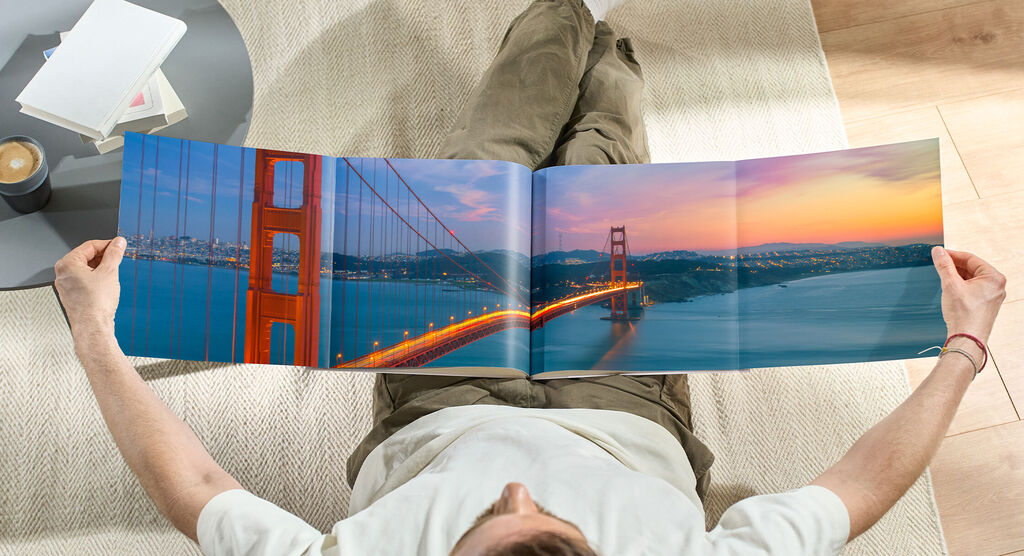 En person sitter bekvämt på golvet och betraktar en öppen fotobok med en vacker bild av Golden Gate-bron vid solnedgång.