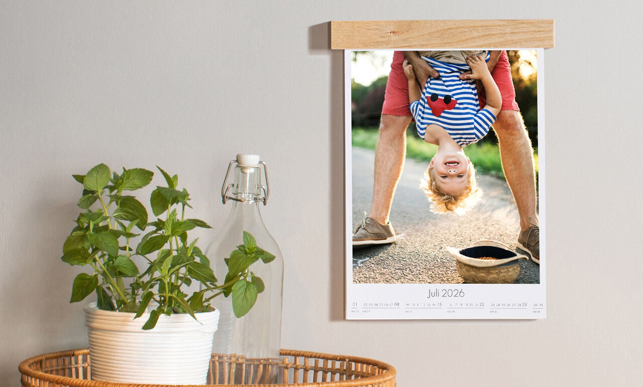 En väggkalender med en glad pojke som står på huvudet, hålls av en vuxen. Bakgrunden visar en solig gata med livsglädje.